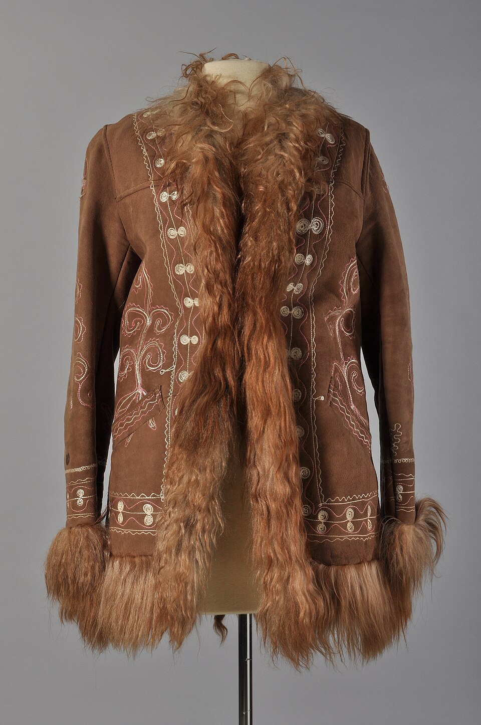Hip-length Afghan coat in light brown suede with long shaggy fleece at the edges and cuffs and cream embroidery down the front, on a museum stand