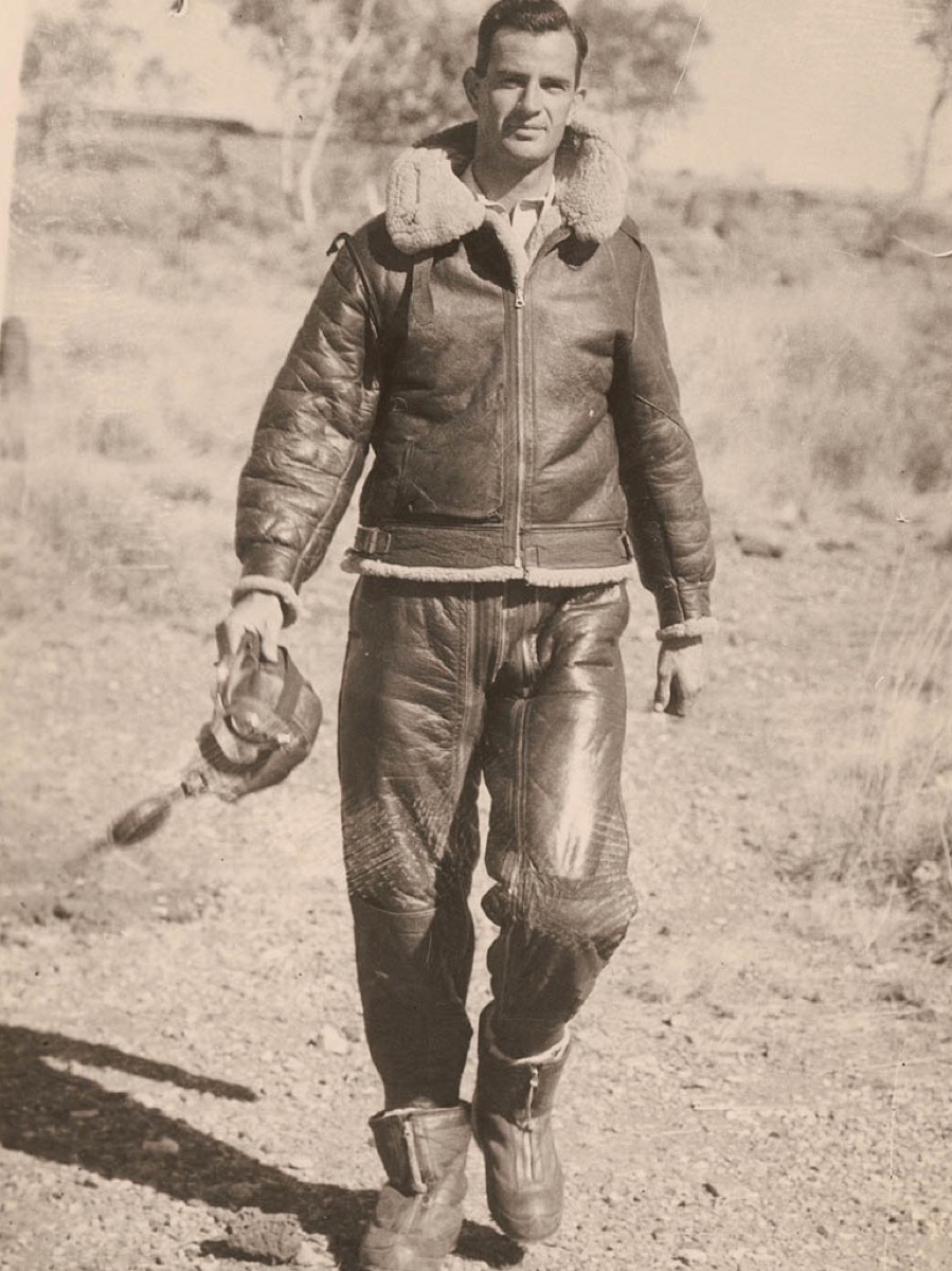 An airman around 1942 in a sheepskin flying jacket with a thick fleece collar and belt, leather flying trousers and fleece-lined boots, holding his flying helmet