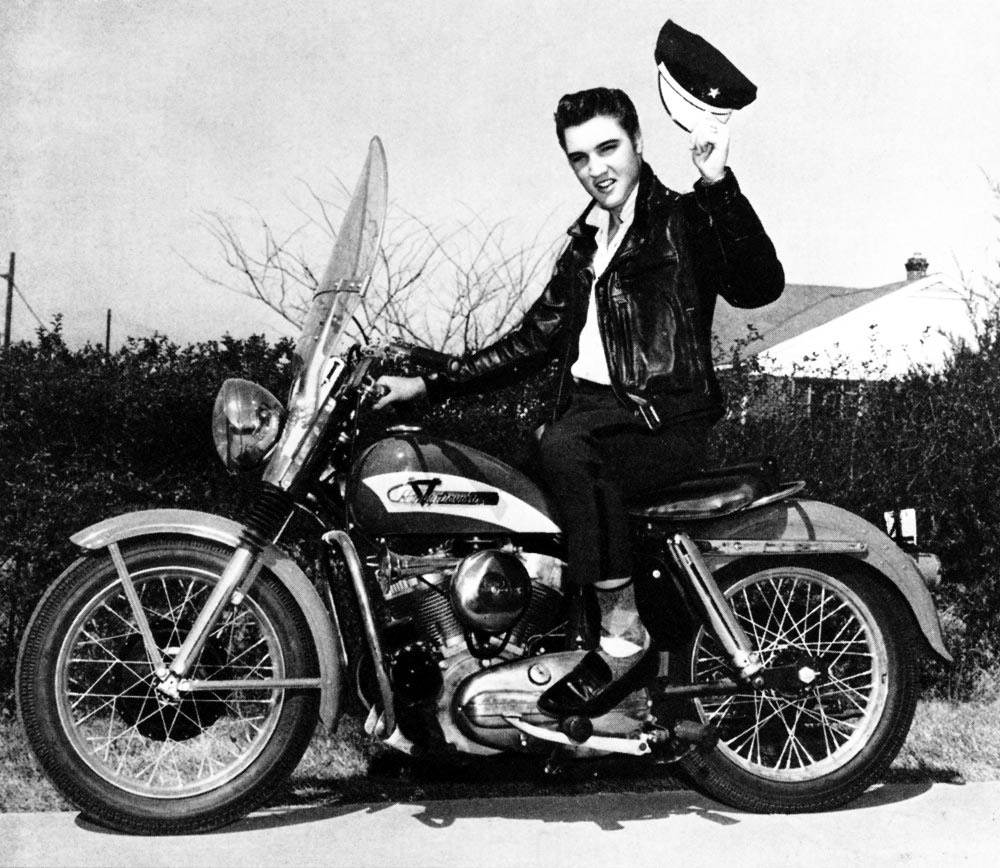 Elvis Presley in 1956 sitting on a Harley-Davidson motorcycle in a black leather jacket, raising his cap
