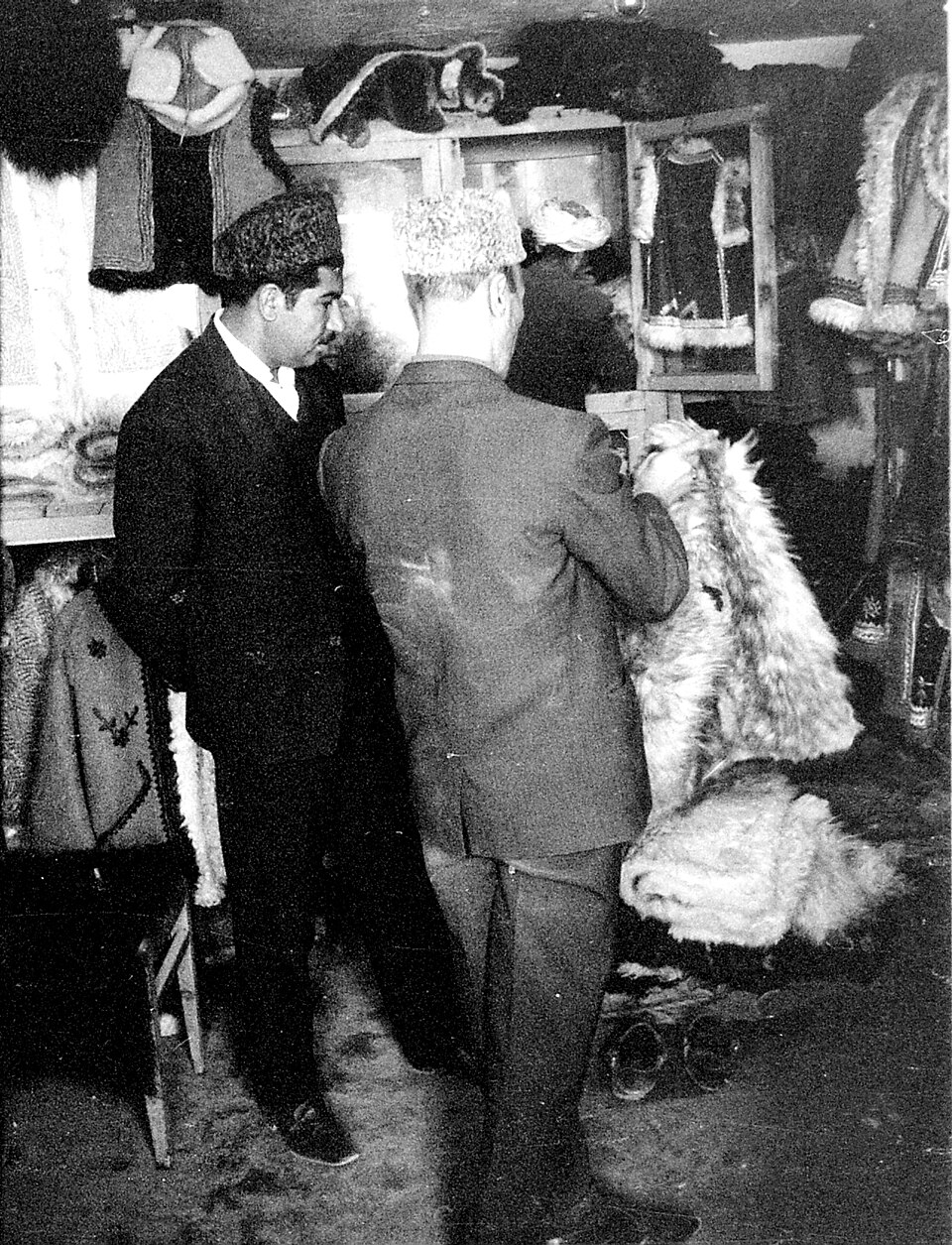 Two men in a fur shop in Kabul in 1966, one holding up a long shaggy sheepskin coat, with embroidered coats hanging on the walls
