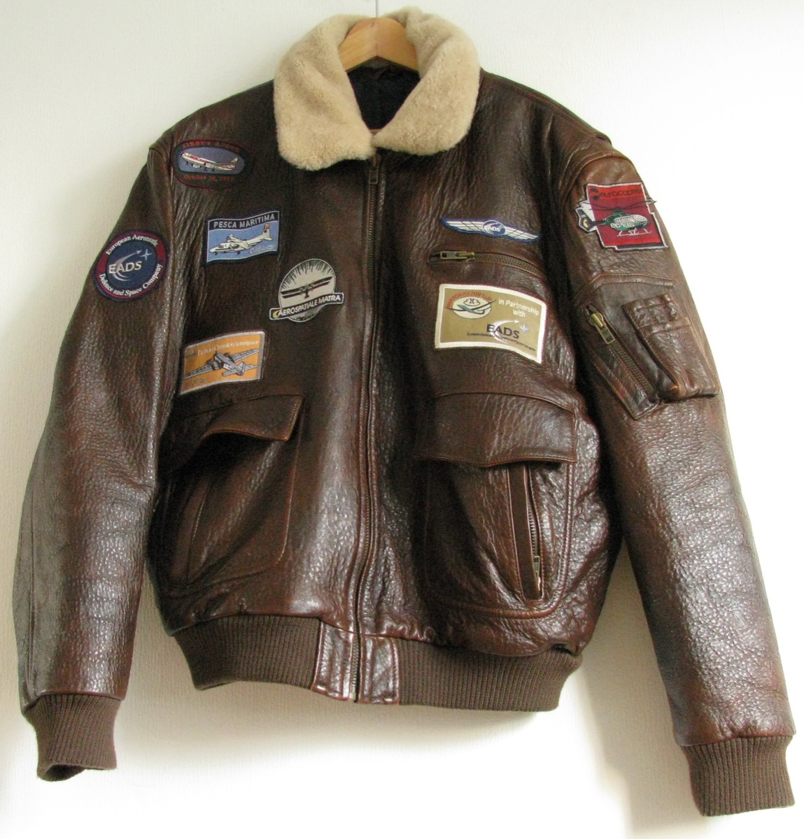 A brown leather flight jacket with a cream shearling collar, ribbed knit cuffs and hem and aviation patches, on a hanger