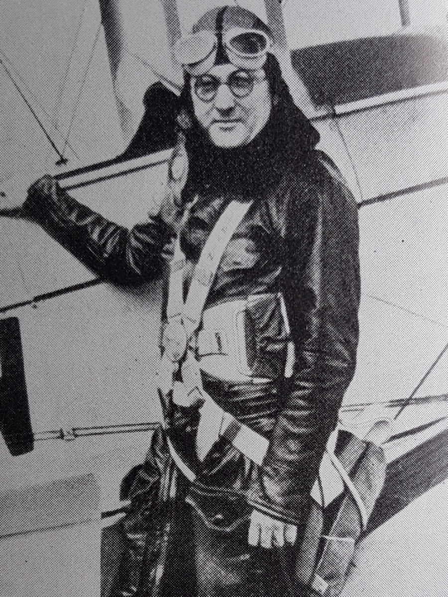 Leslie Irvin in leather flying kit, helmet and goggles, wearing a parachute harness, beside a biplane