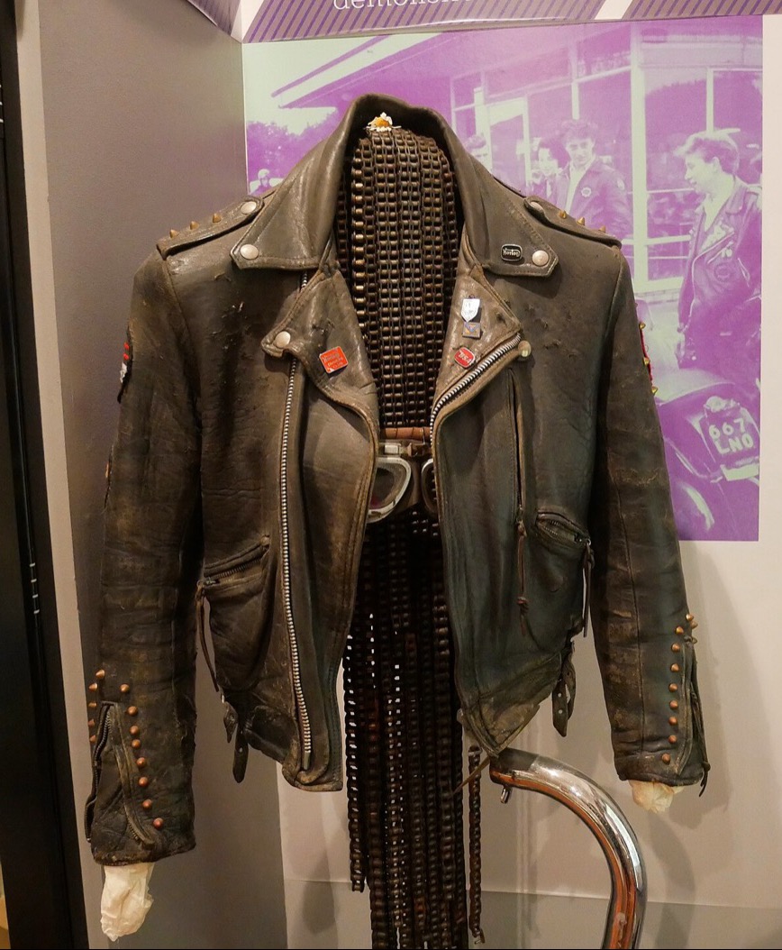 A worn black 1950s leather biker jacket decorated with studs and badges, displayed in a museum case